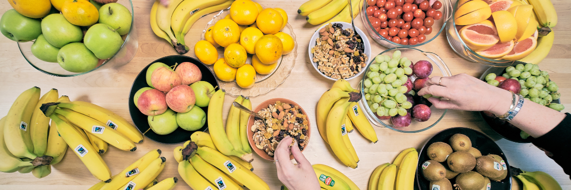 Pöydällä on erilaisia hedelmiä, kuten banaaneja, omenoita ja kiivejä. Kuvassa näkyy myös käsiä, jotka ottavat hedelmiä.